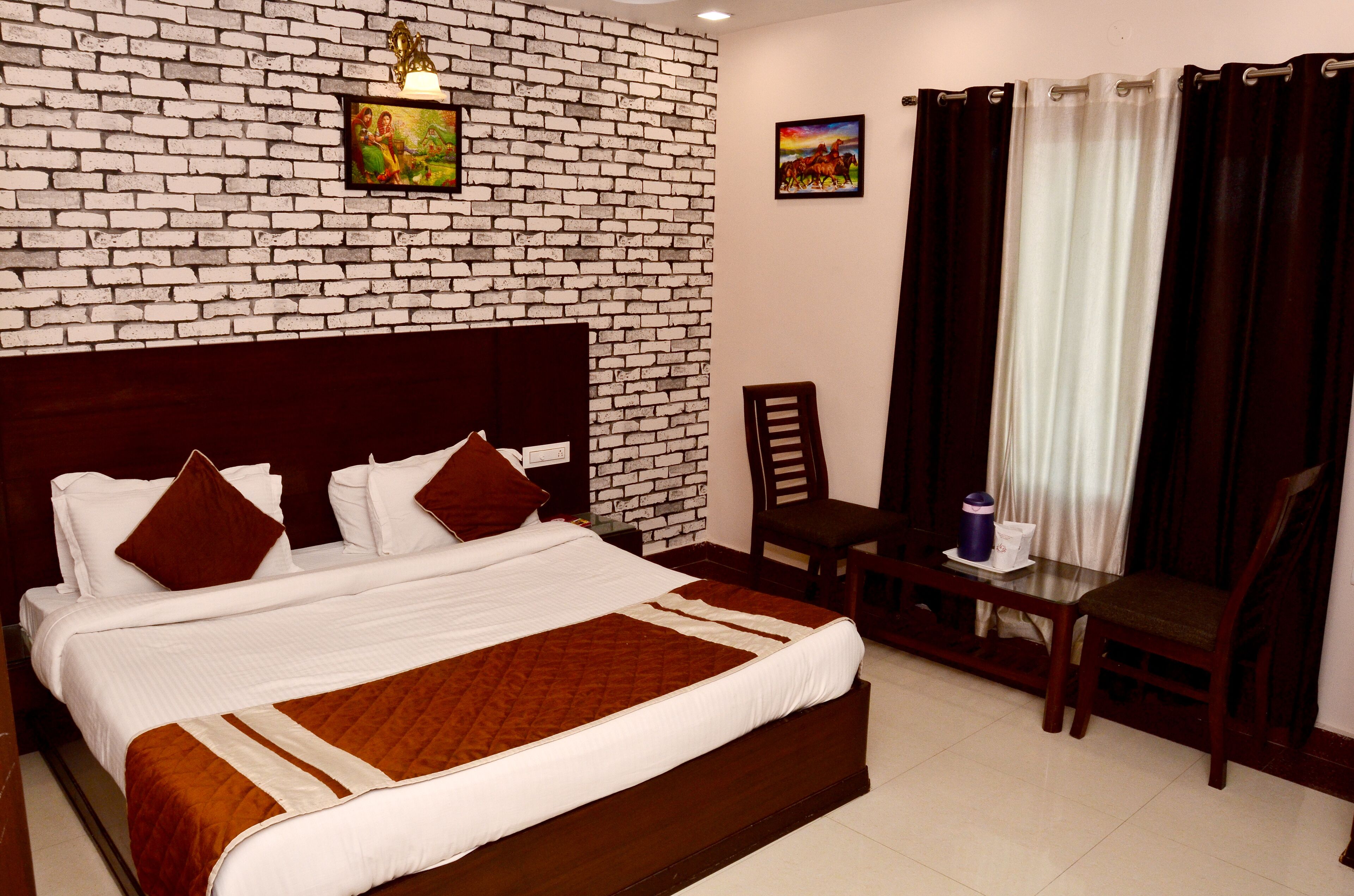 A hotel room with a brick-patterned wall, a large bed with white bedding and brown accents, a small table with a chair, and curtains on the window.