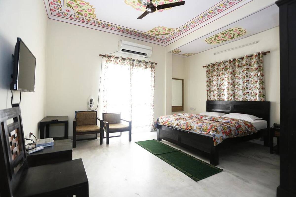 A bedroom with a bed, a chair, a desk, a television, and an air conditioning unit.