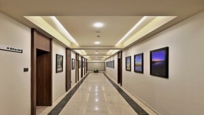 A long, well-lit hallway with framed pictures on the walls, a sign indicating room numbers, and a polished floor. - Vits Devbhumi Hotel (Dwarka)