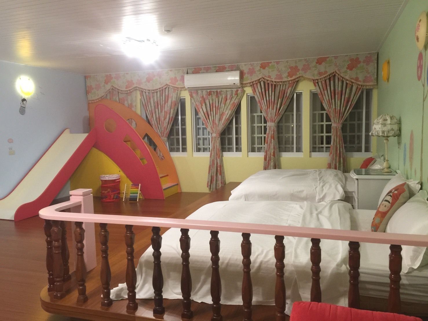 A bedroom with a bed, a wooden railing, a window with curtains, and a small table with a lamp.