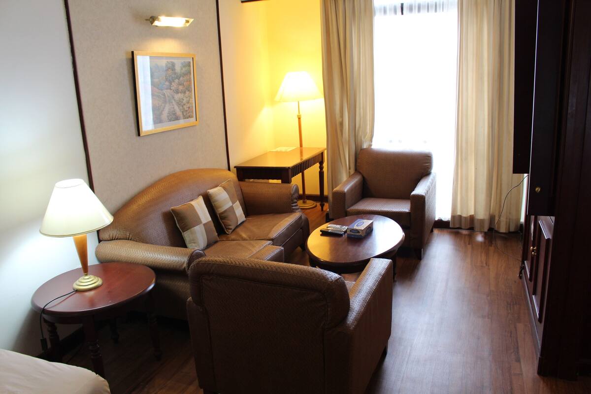 A hotel room with a sofa, armchair, and a small table with a lamp.