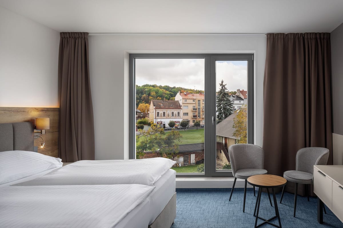 A hotel room with a large window, a bed, a desk, two chairs, and a lamp.
