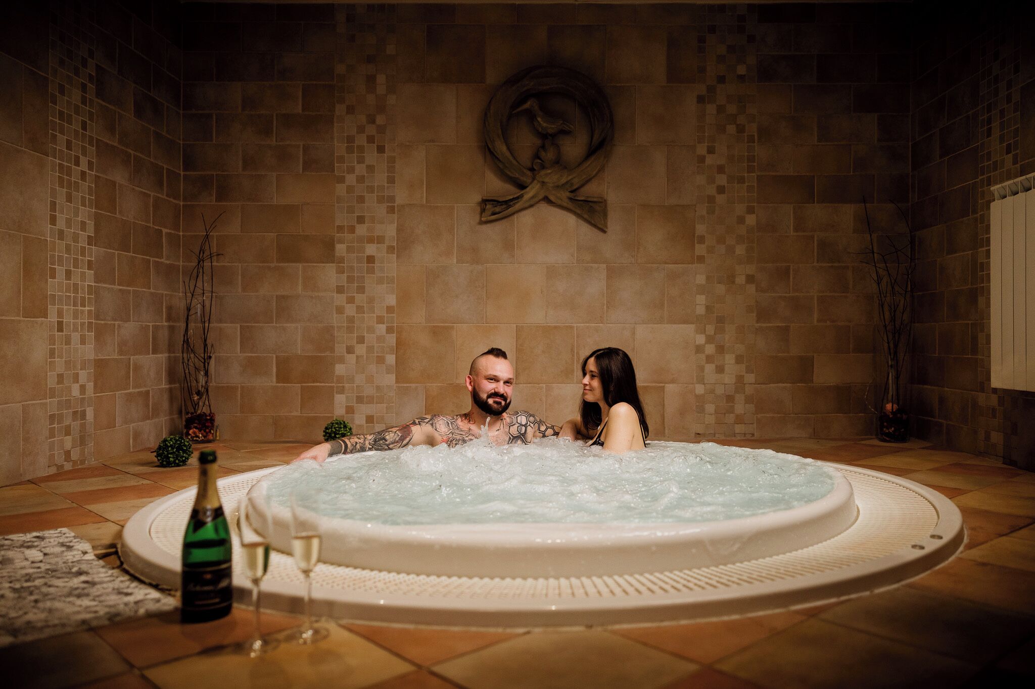Un couple dans un jacuzzi, avec une bouteille de champagne et des verres posés sur le sol.