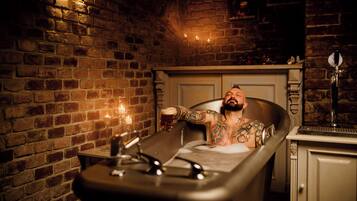 A person relaxing in a bathtub with a brick wall and a shelf with bottles in the background.