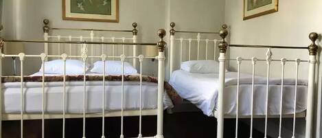 Una habitación con dos camas, cada una con ropa de cama blanca y estructura metálica blanca. Hay dos cuadros en las paredes.