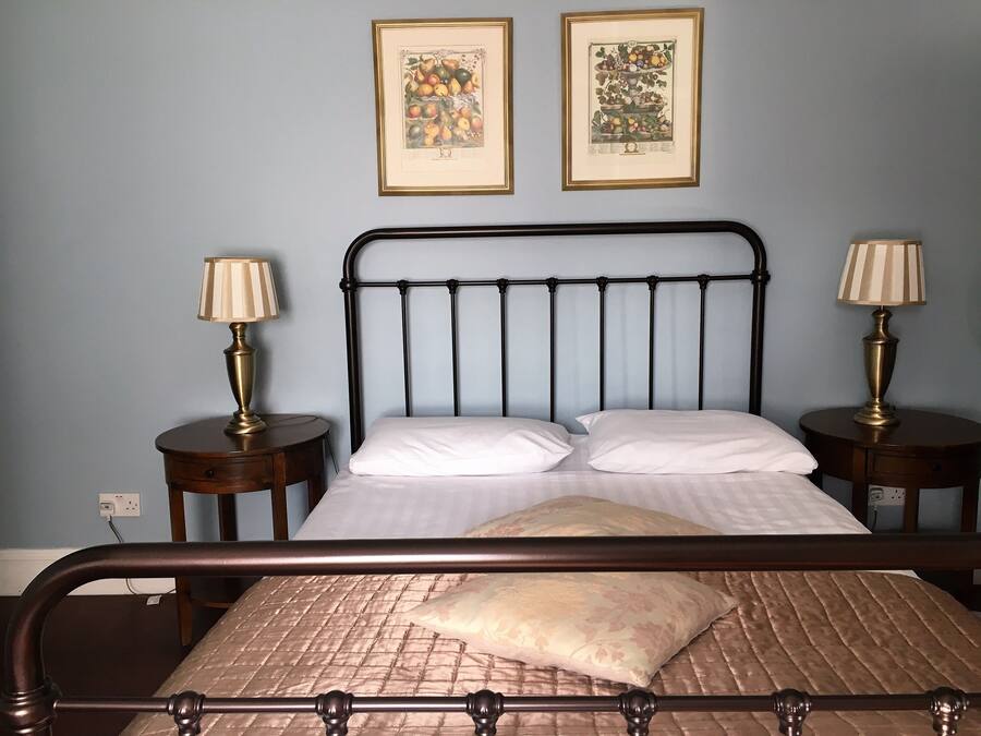 Una cama bien tendida con ropa de cama blanca y una colcha acolchada, flanqueada por dos mesitas de noche con lámparas, frente a una pared azul claro.