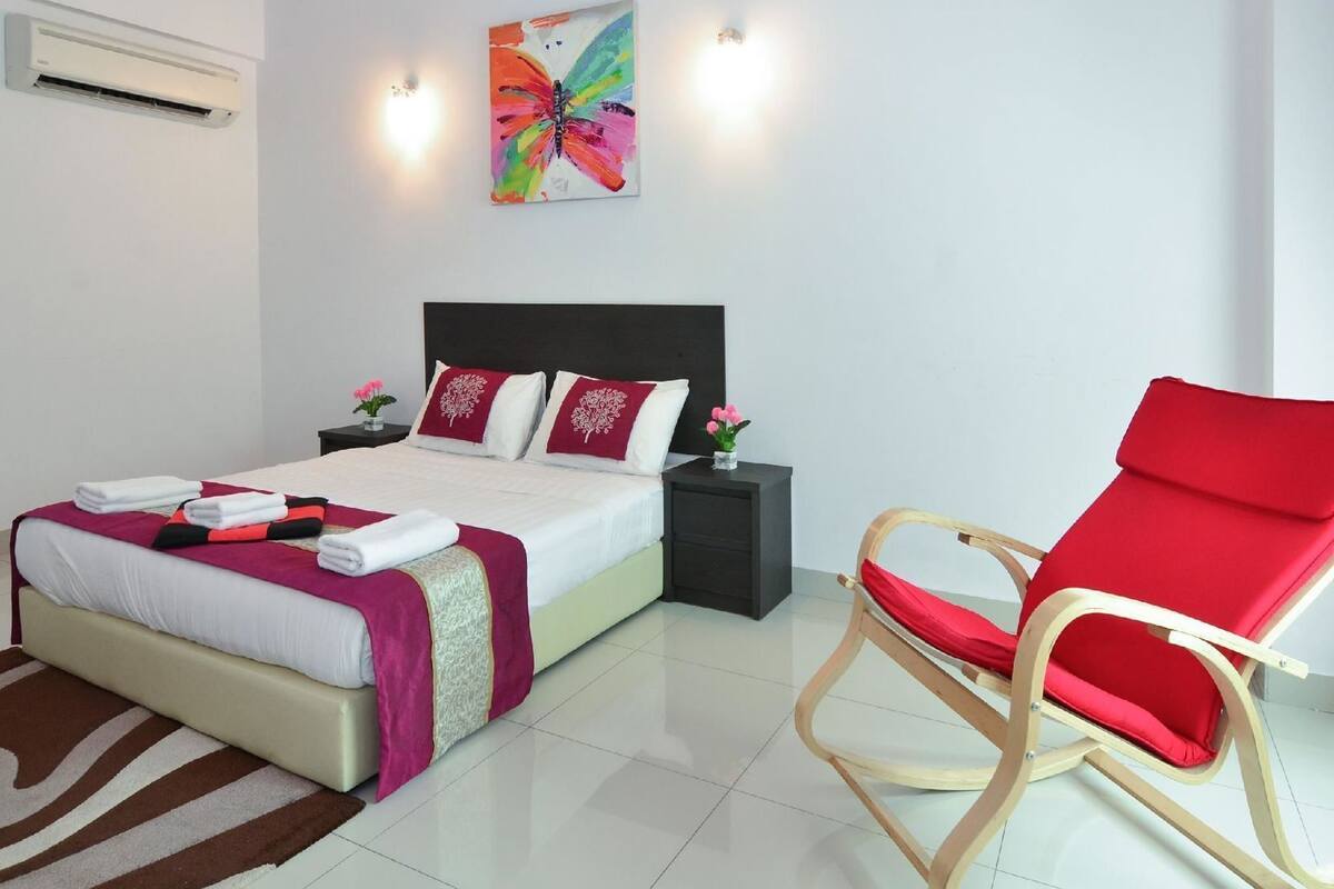 Una habitación de hotel con cama, una butaca roja, una mesita de noche, un aire acondicionado de pared y un cuadro de colores.
