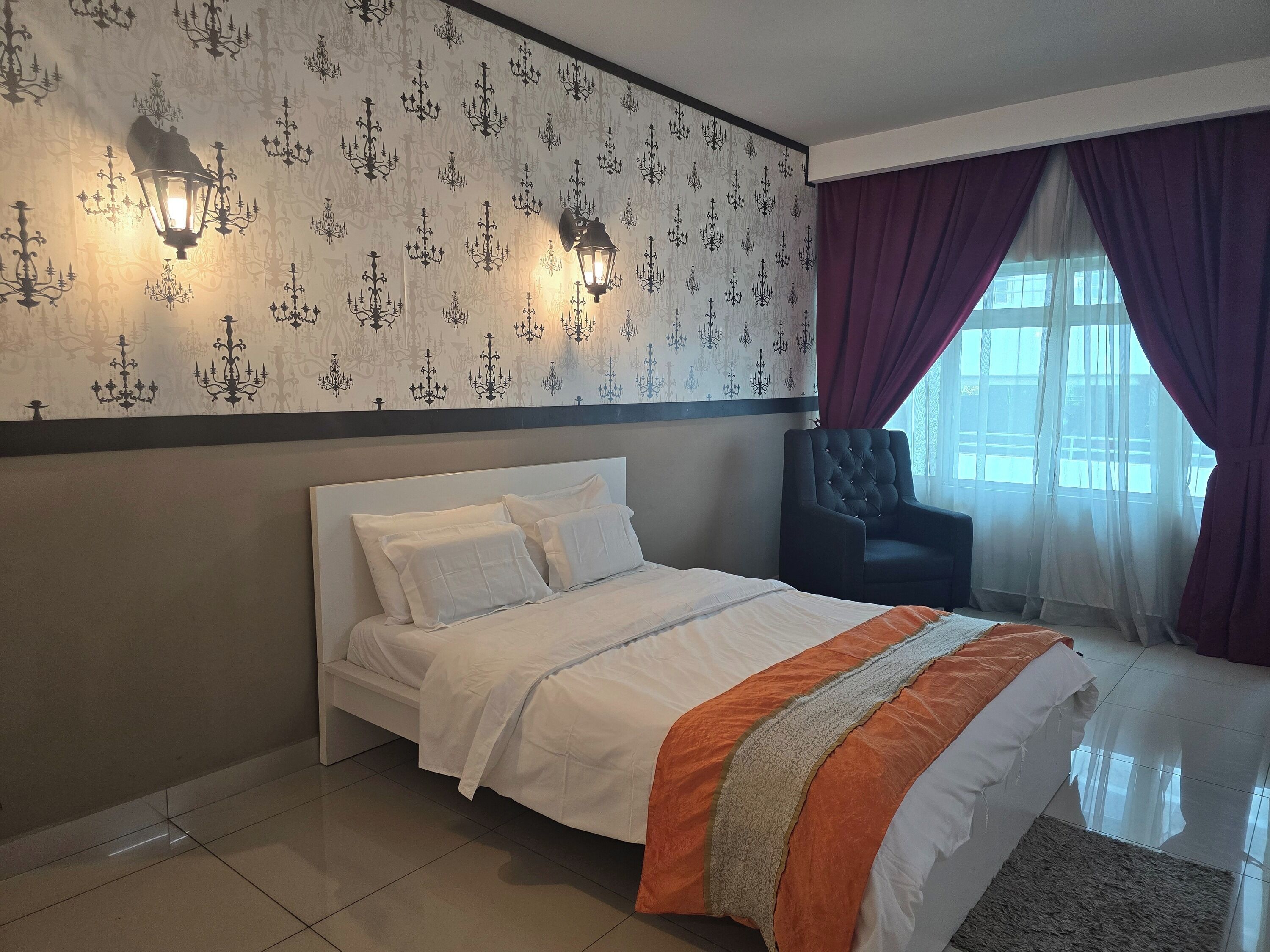 A neatly made bed with white bedding and an orange accent, a black upholstered armchair, and a wall with a chandelier pattern.
