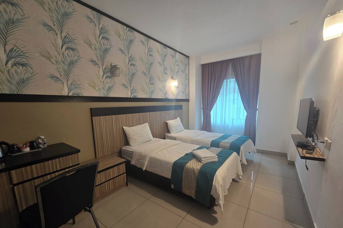 Habitación de hotel con dos camas, un escritorio, una silla, un televisor y un ventanal grande con cortinas.