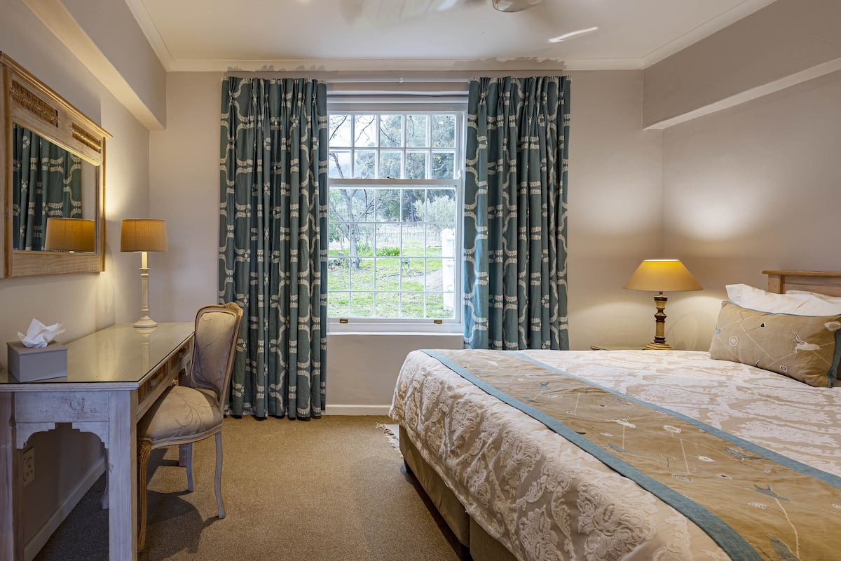 A hotel room with a bed, two bedside lamps, a desk with a chair, a mirror, and a window with curtains.