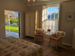 A bedroom with a bed, two wicker chairs, a small table with a vase, and a view of mountains through a window. - L'Auberge Chanteclair (Franschhoek)
