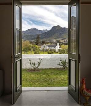 An open door leading to a view of a rural landscape with mountains, a white building, and a fenced area with small trees. - L'Auberge Chanteclair (Franschhoek)