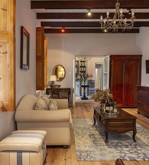 A living room with a sofa, a coffee table with wine bottles, a chandelier, wooden furniture, and a bookshelf in the background. - L'Auberge Chanteclair (Franschhoek)