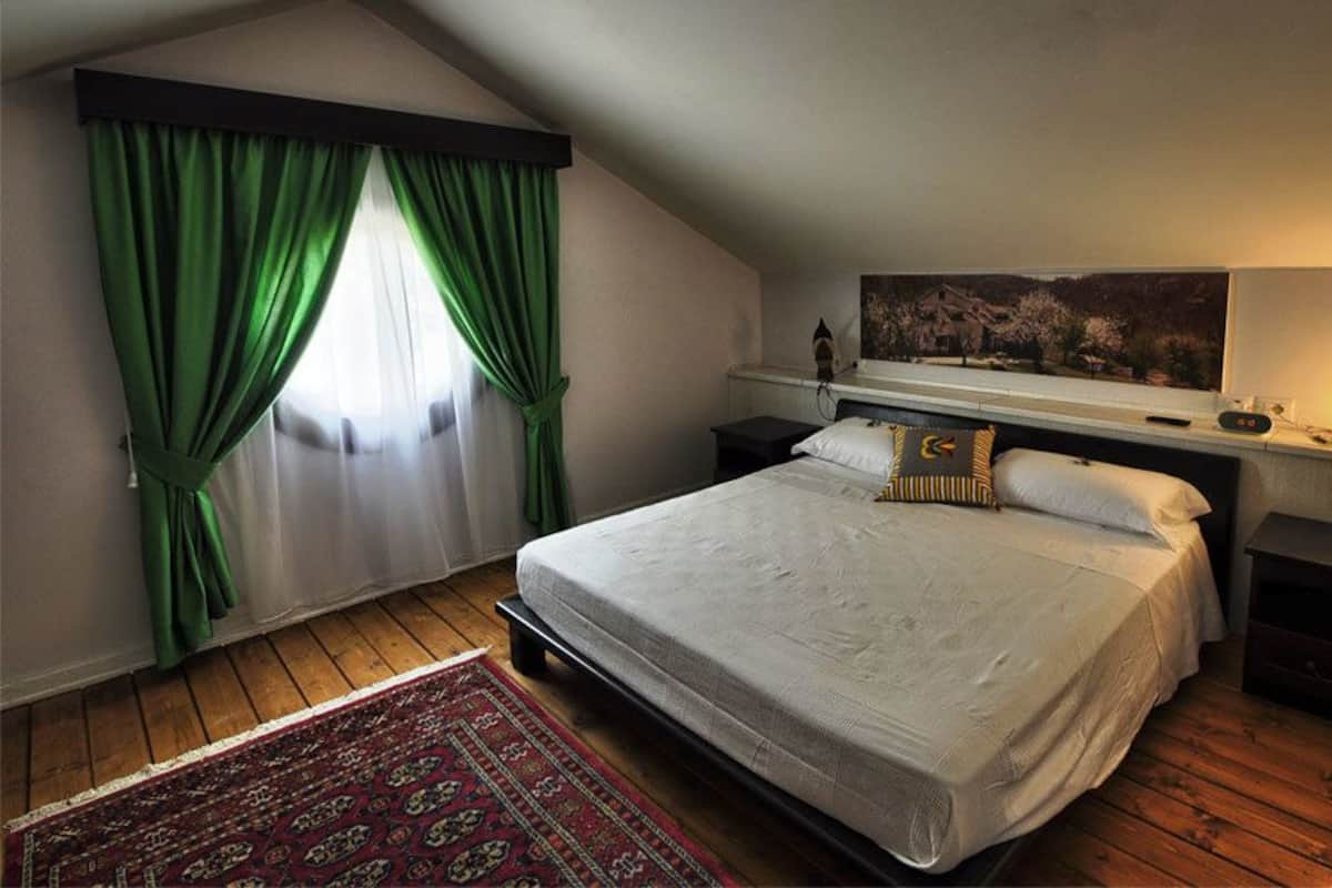 A bedroom with a large bed, green curtains, a painting above the bed, a bedside table with a lamp, and a fireplace.