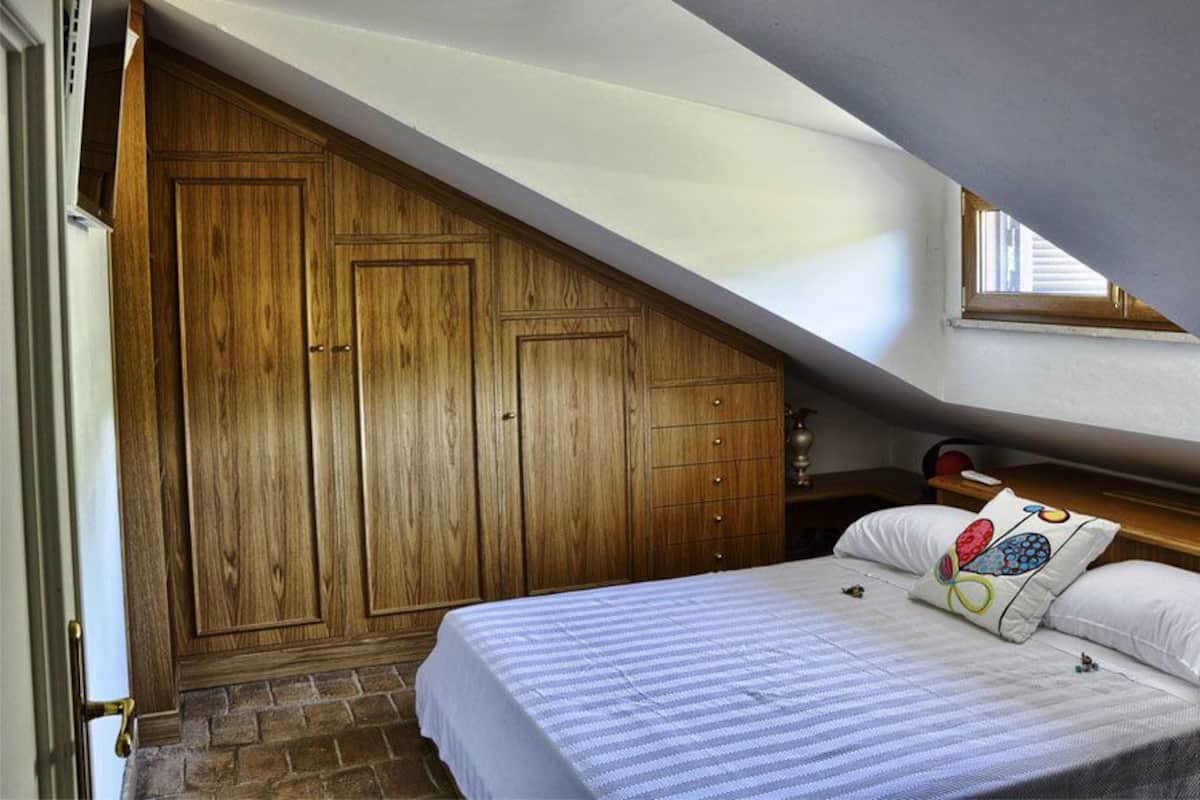 A bedroom with a sloped ceiling, wooden wardrobe, and a bed with patterned pillows.