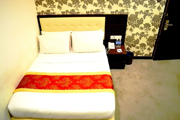Un lit bien fait, avec du linge de lit blanc et une couvre-lit rouge et or, une table de chevet avec un téléphone et une bouteille, et un papier peint à motifs floraux sur le mur.