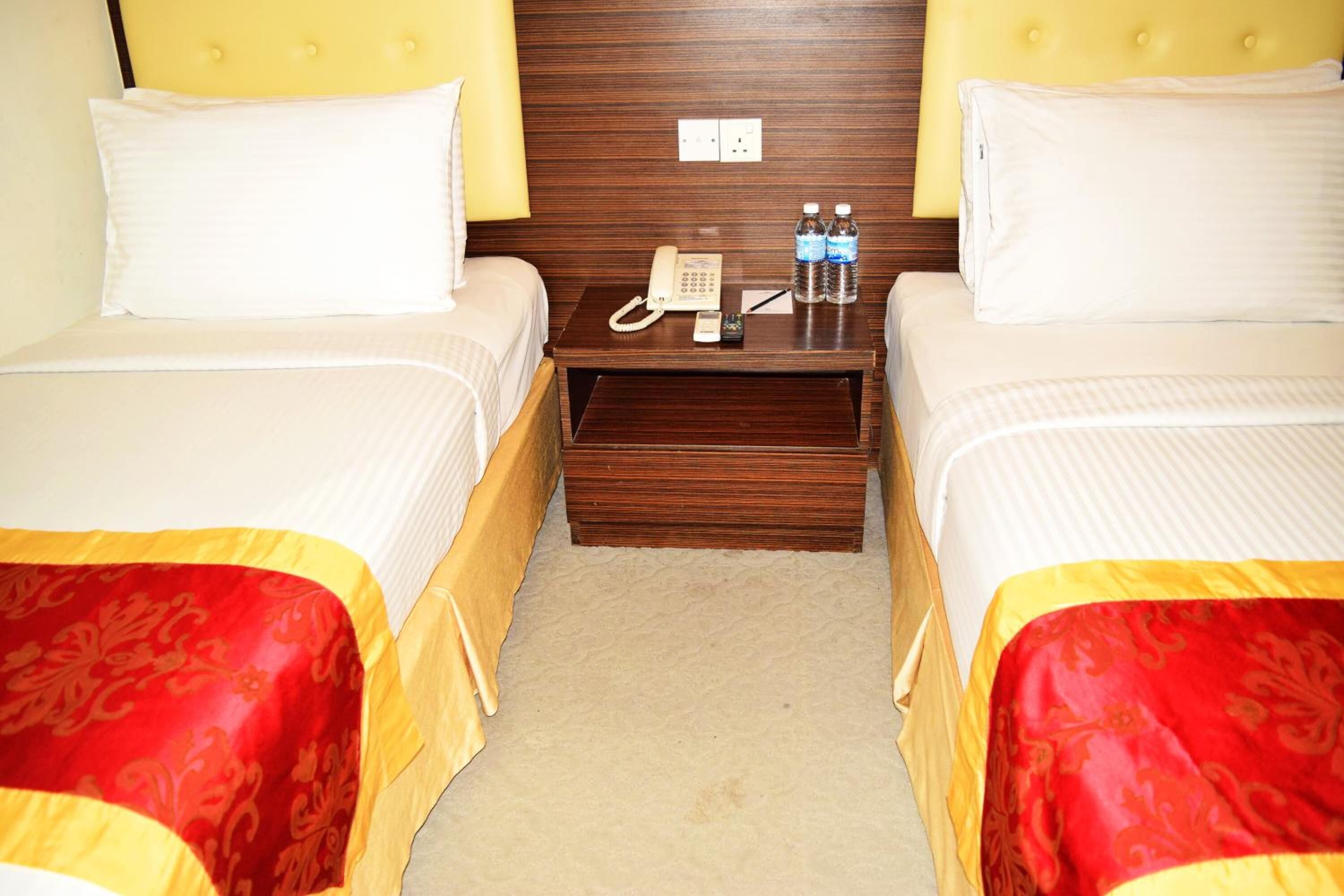 A hotel room with two beds, a wooden headboard, a nightstand with a phone and water bottles, and a carpeted floor.