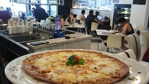 Une grande pizza servie sur une assiette blanche dans un restaurant, avec des clients qui dînent en arrière-plan.