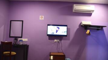 A room with purple walls, a television mounted on the wall, a desk with a chair, a trash bin, and an air conditioning unit.