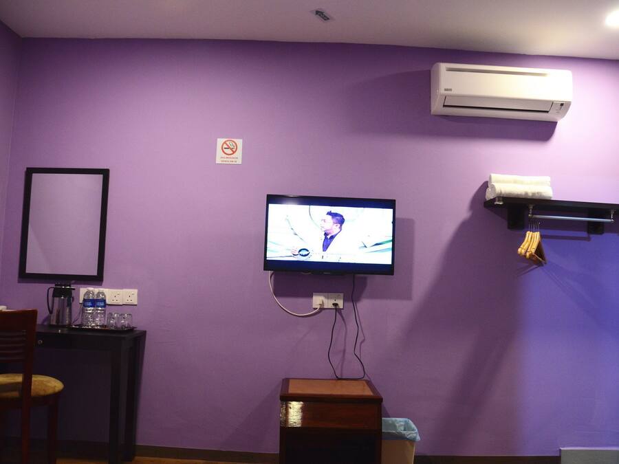 A room with purple walls, a television mounted on the wall, a desk with a chair, a trash bin, and an air conditioning unit.