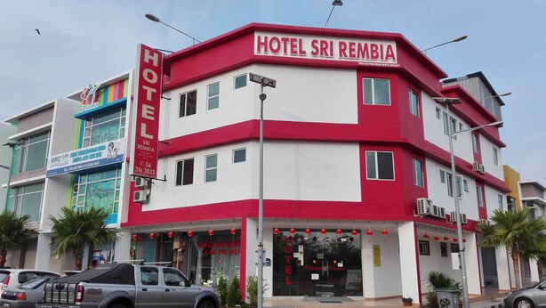Un immeuble à plusieurs étages, dont la façade se distingue par un contraste marqué entre le rouge et le blanc, avec une enseigne indiquant «Hôtel Sri Rempia» et une entrée bien visible, dotée d’une porte en verre.