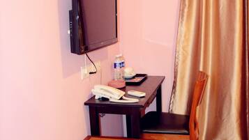 A small room with a television mounted on the wall, a wooden table with a chair, a small cabinet, and a curtain.