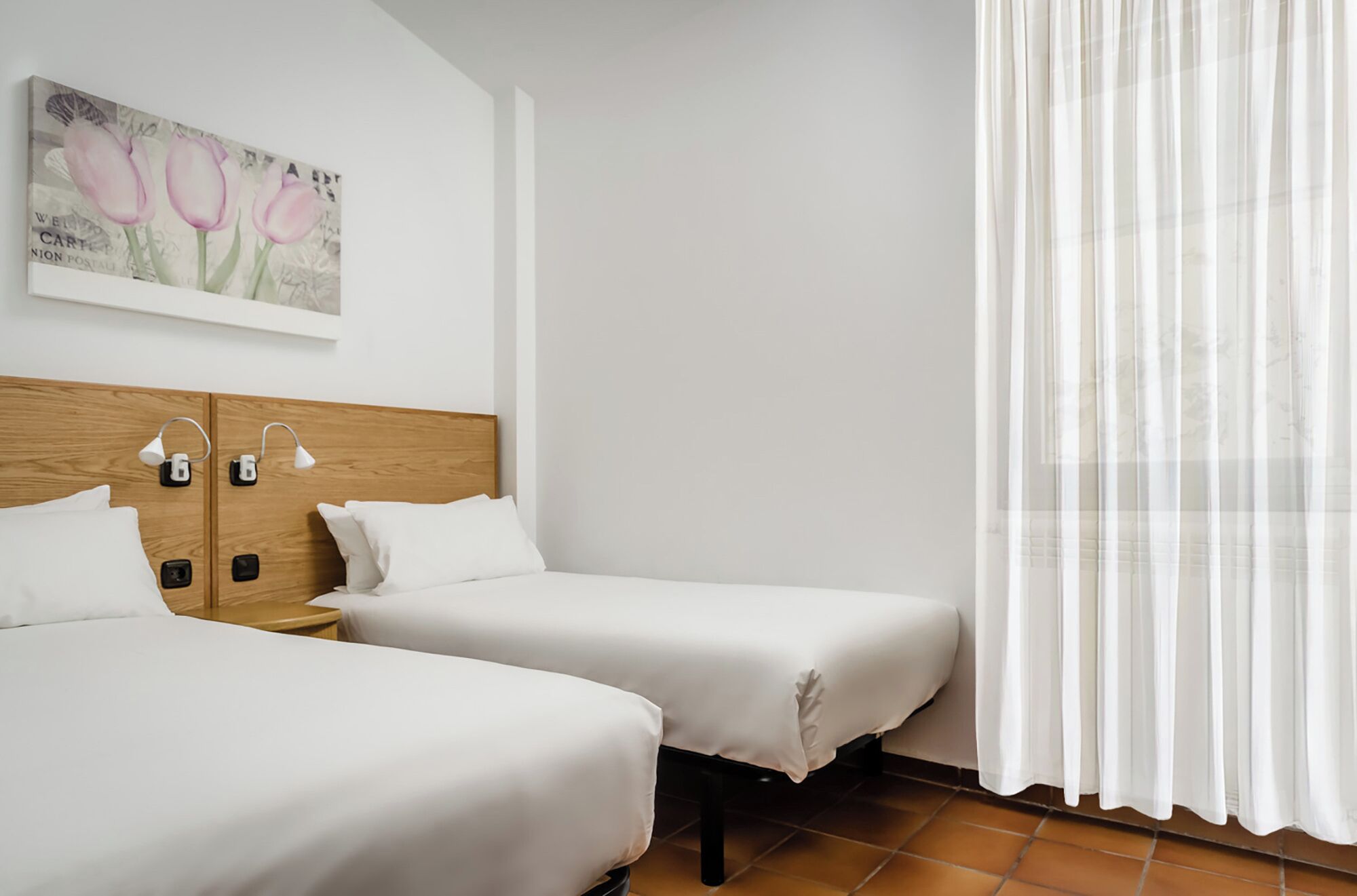 Une chambre d’hôtel avec deux lits simples, une tête de lit en bois, un tableau représentant des tulipes, une fenêtre avec des rideaux en voile et un sol carrelé.