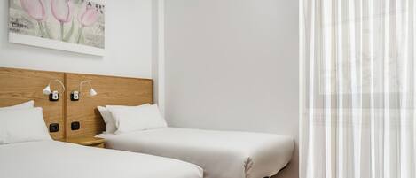 A hotel room with two single beds, a wooden headboard, a painting of tulips, a window with sheer curtains, and a tiled floor.