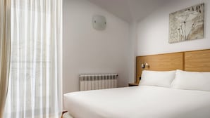 A bedroom with a large bed, a radiator, a wall-mounted light, and a window with sheer curtains.