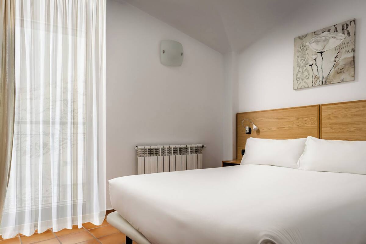 A bedroom with a large bed, a radiator, a wall-mounted light, and a window with sheer curtains.