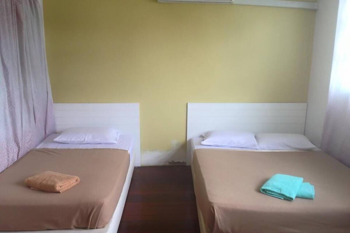 Une chambre avec deux lits, chacun doté d’une tête de lit blanche et d’une couverture de lit marron. Il y a une fenêtre sur le côté droit, et un appareil de climatisation fixé au mur au-dessus des lits.