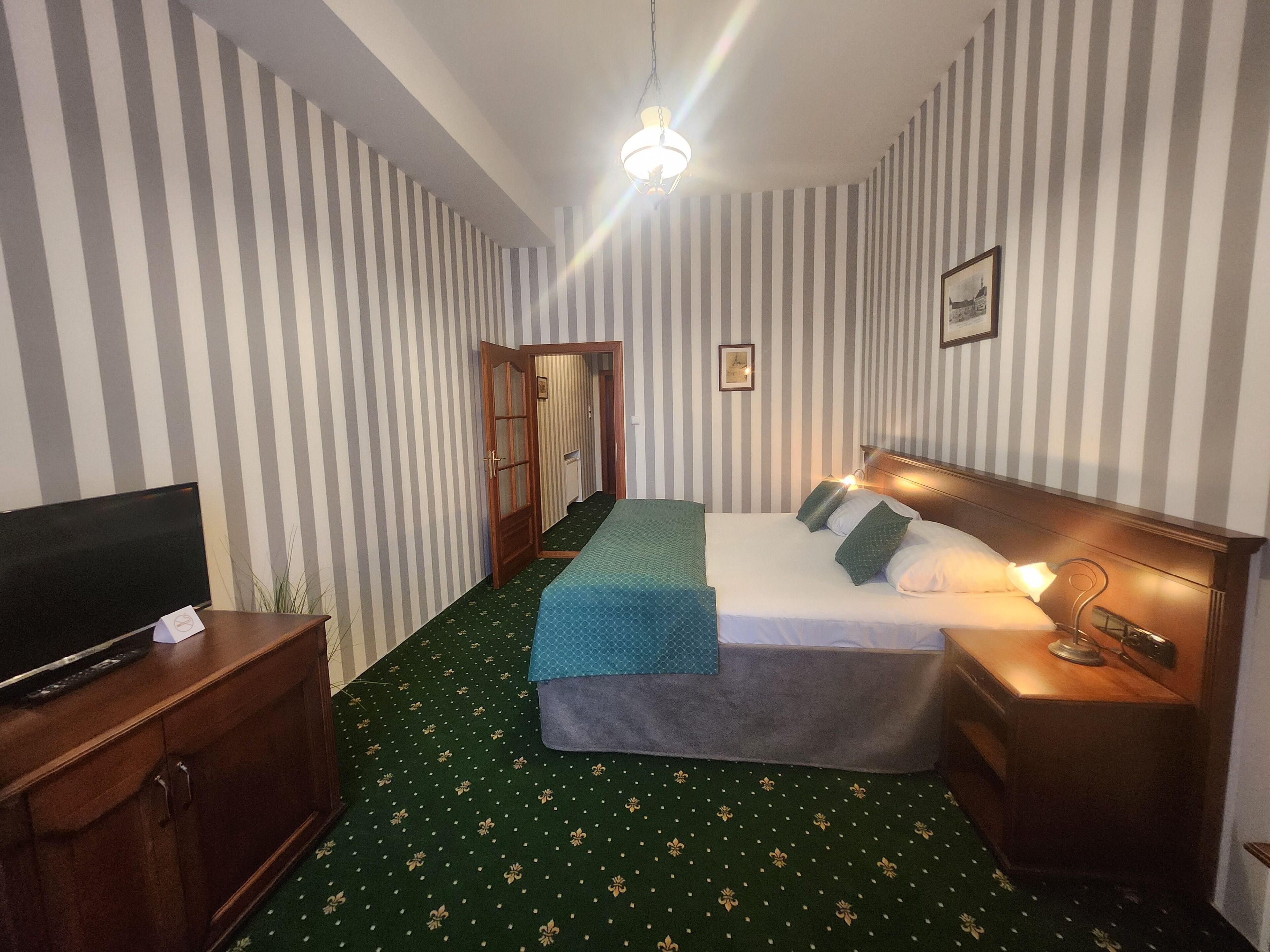 Habitación de hotel con papel tapiz a rayas, una cama con almohadas, un televisor en un mueble de madera y una alfombra verde.