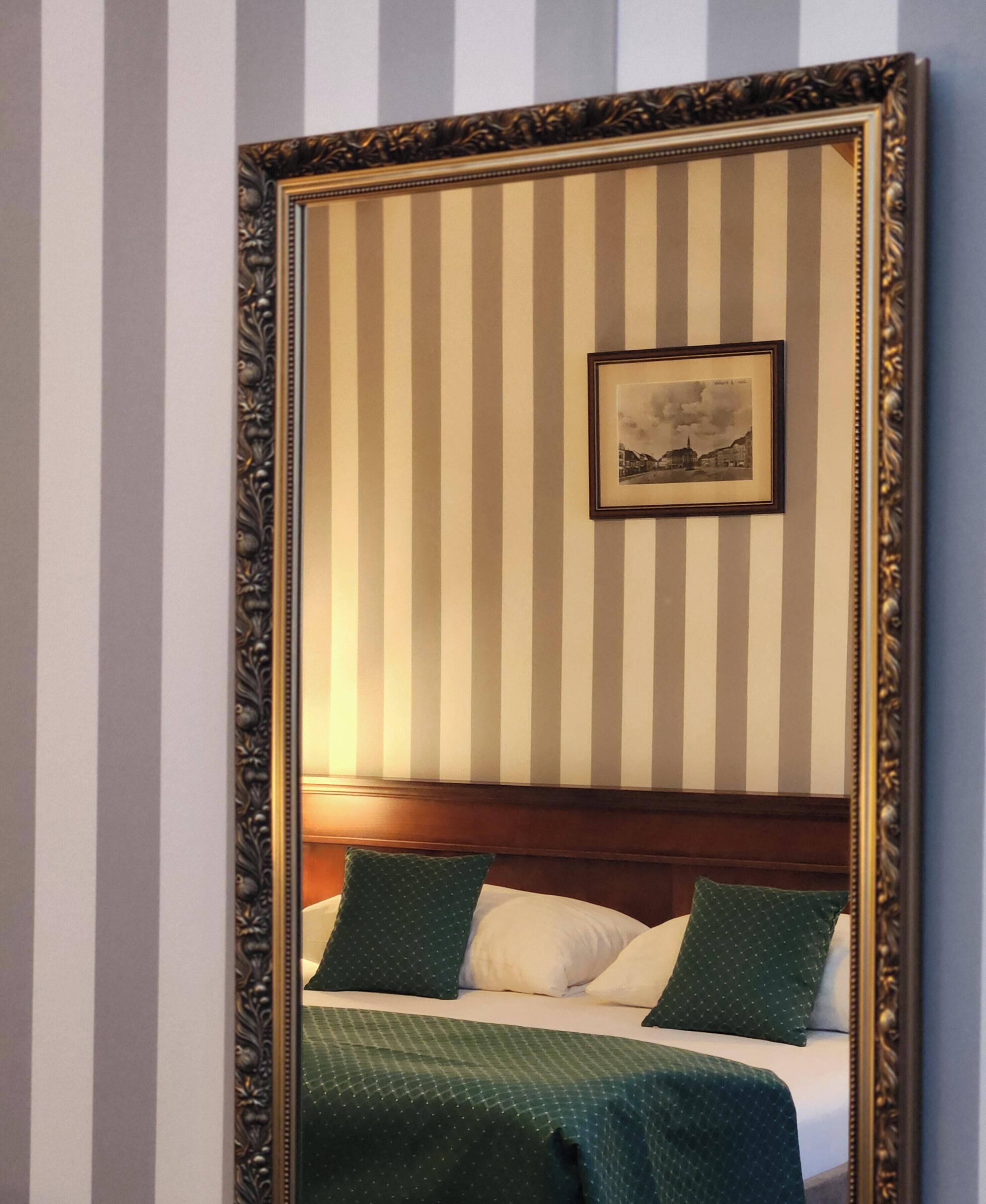 Ein Zimmer mit einem Bett, einem Kopfteil, einem gerahmten Bild an der Wand und einem Spiegel mit aufwendiger Rahmenornierung.