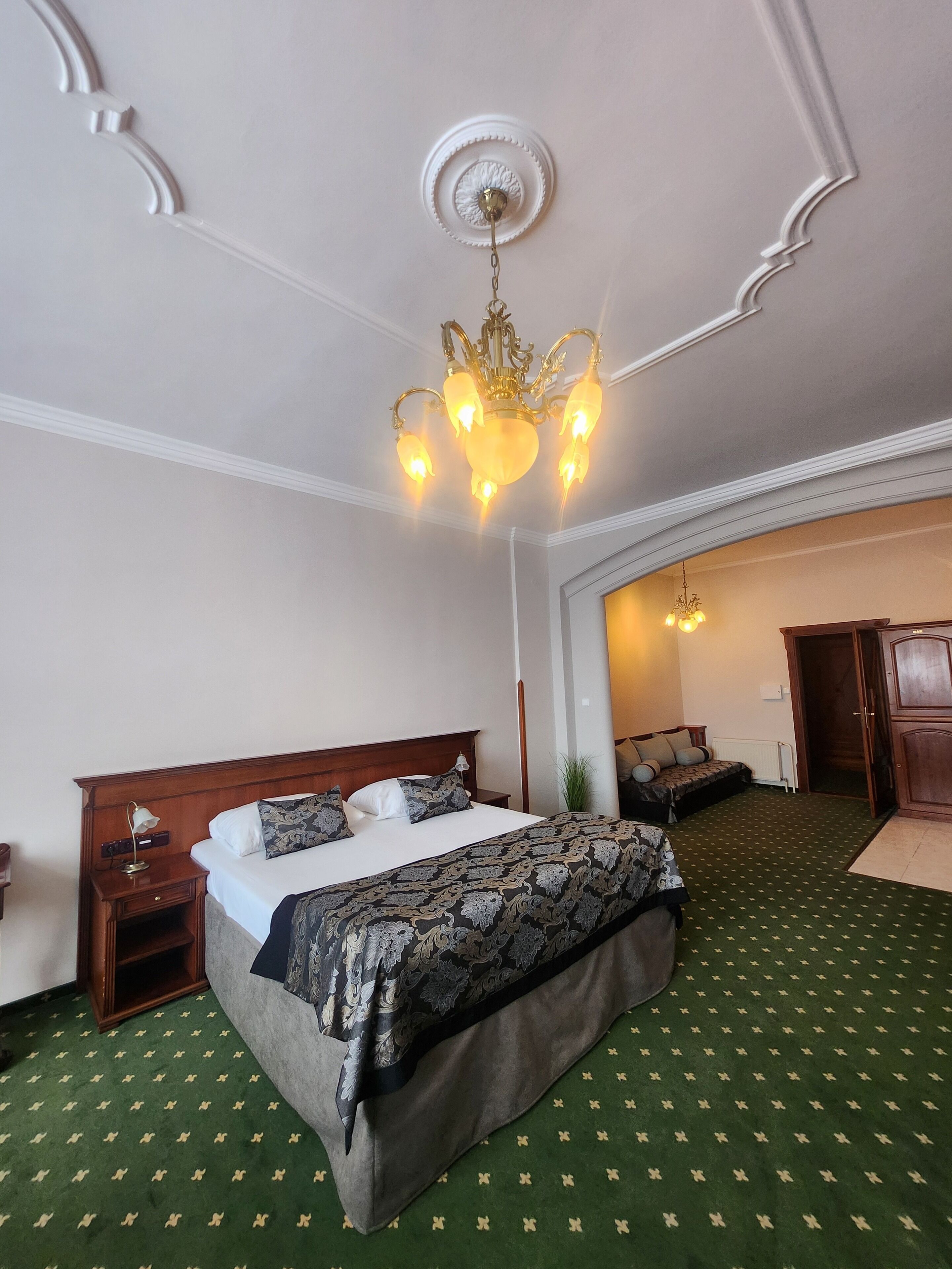 A hotel room with a large bed, a wooden headboard, a nightstand, a lamp, a green patterned carpet, and a chandelier.