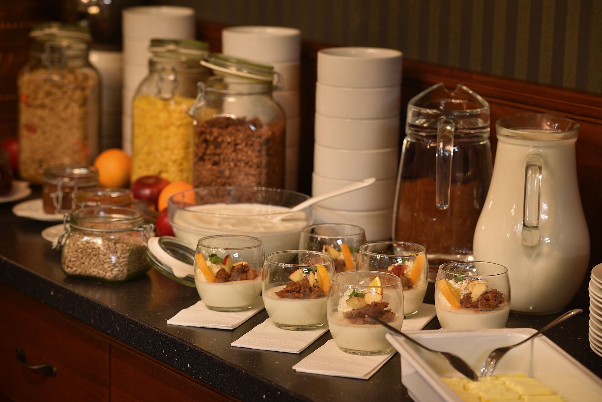 Una mesa de buffet con diversos alimentos, incluyendo frascos de conservas, un tazón de sopa y varios vasos de postre.