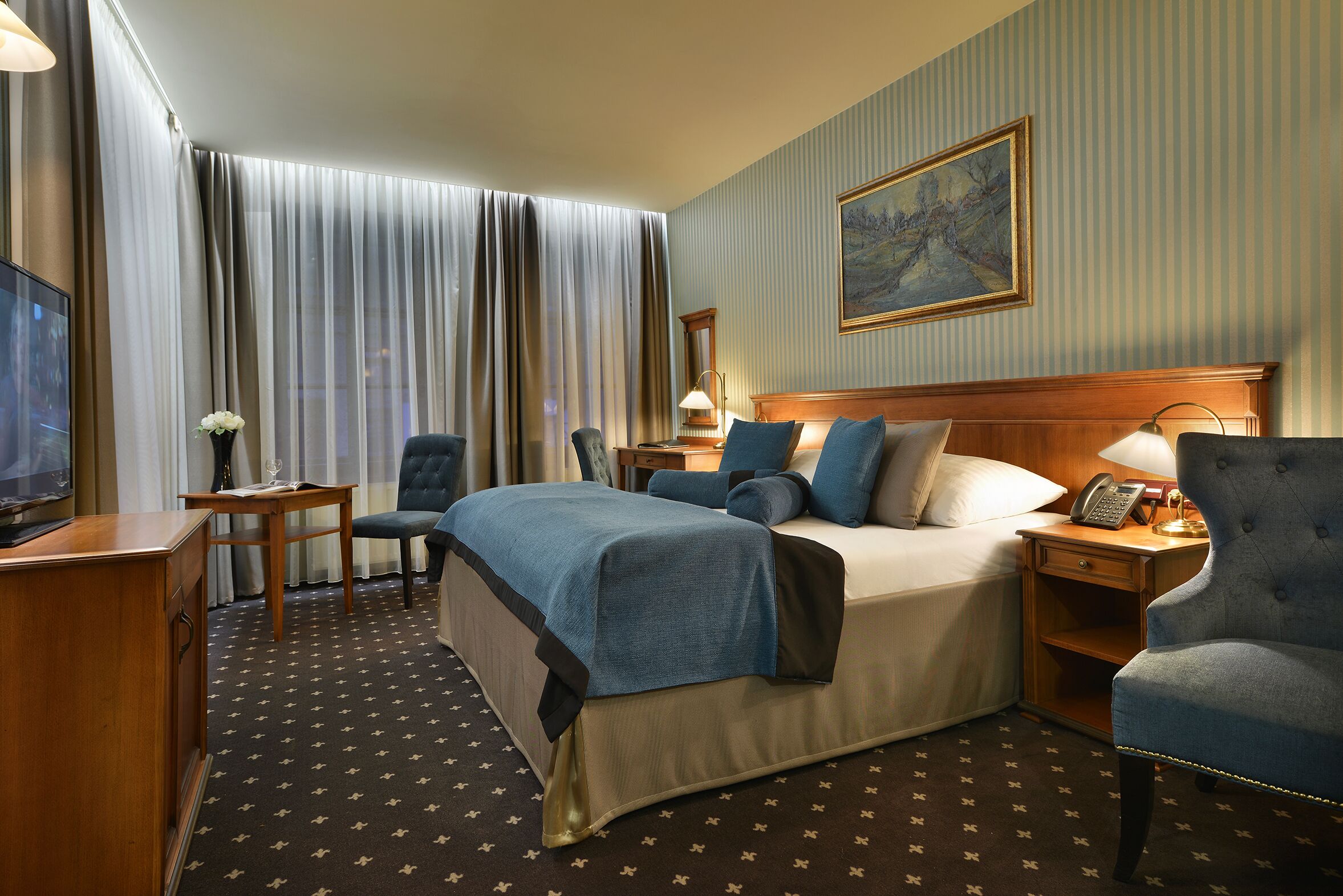 Een hotelkamer met een bed, twee fauteuils, een bureau met lamp, een televisie en een schilderij aan de muur.