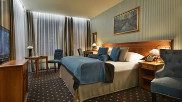 Een hotelkamer met een bed, twee fauteuils, een bureau met lamp, een televisie en een schilderij aan de muur.