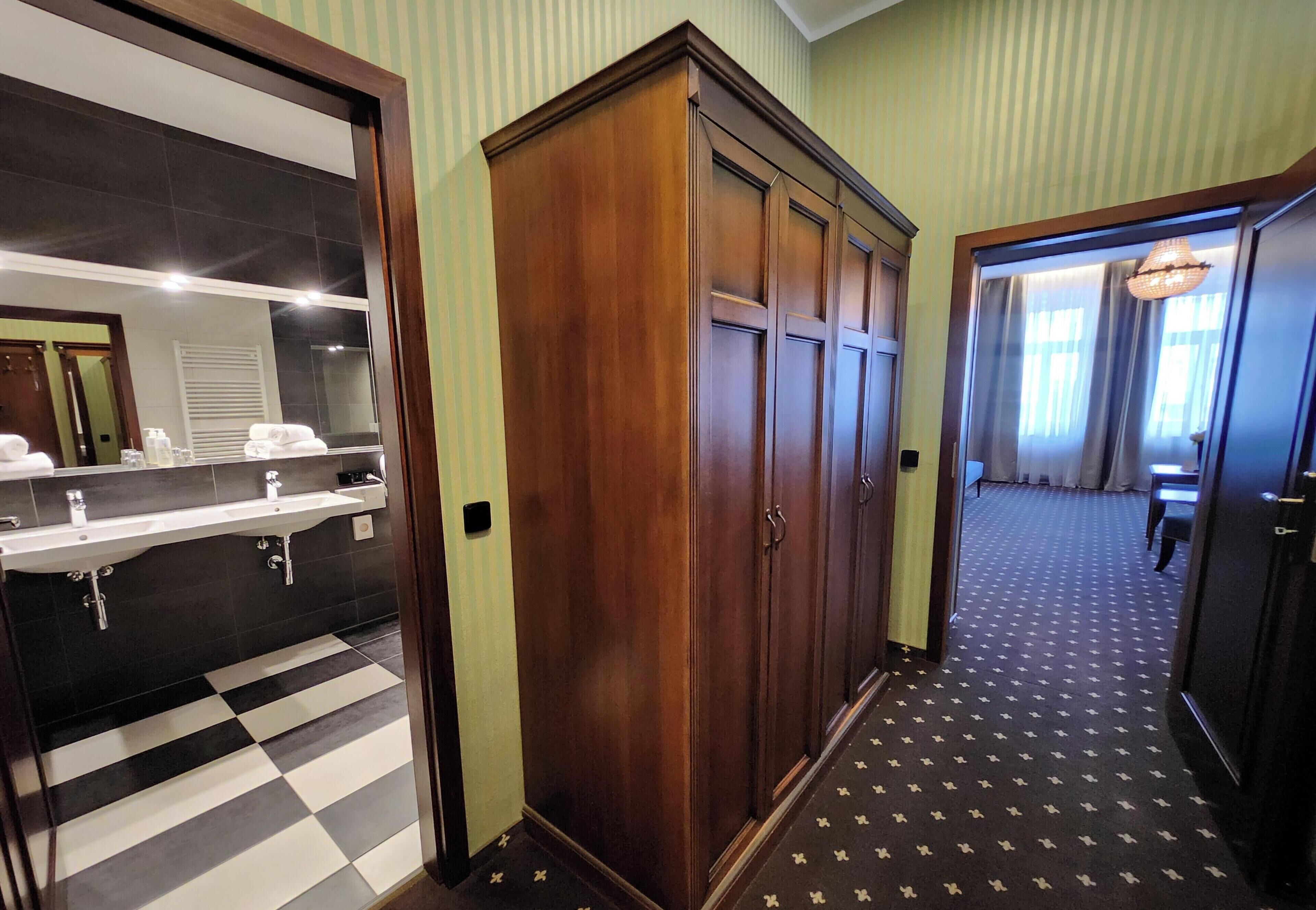 A hotel hallway with a checkered floor, a wooden wardrobe, a mirror, and a sink.