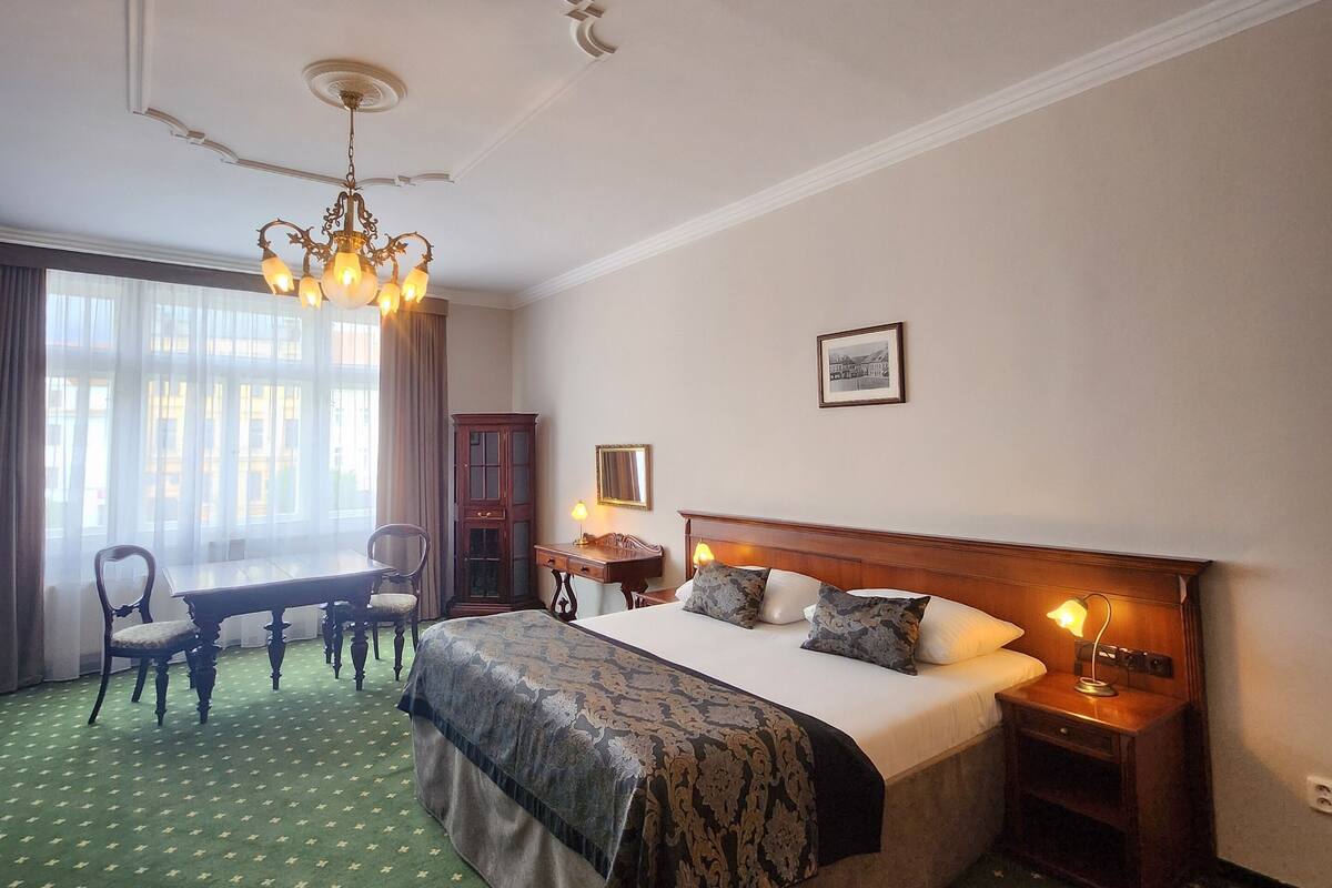 Hotelkamer met een groot bed, een kroonluchter, een eettafel met stoelen en uitzicht op de stad door grote ramen.