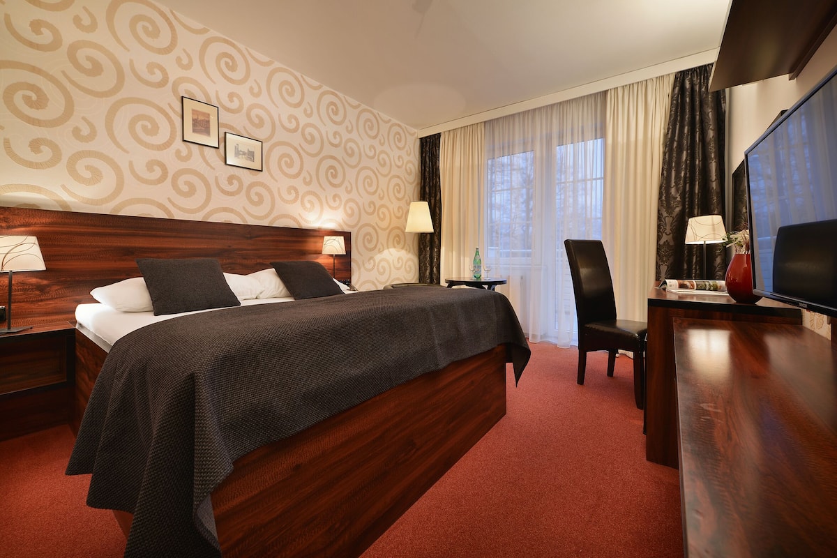Quarto de hotel com uma cama grande, uma mesa de trabalho com cadeira, televisão e janela com cortinas.