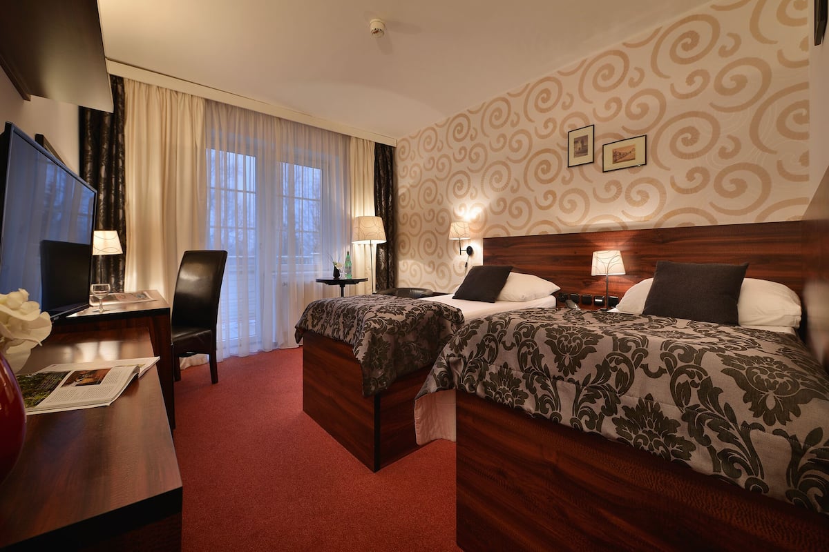 Quarto de hotel com duas camas, uma escrivaninha, televisão e janela com cortinas.