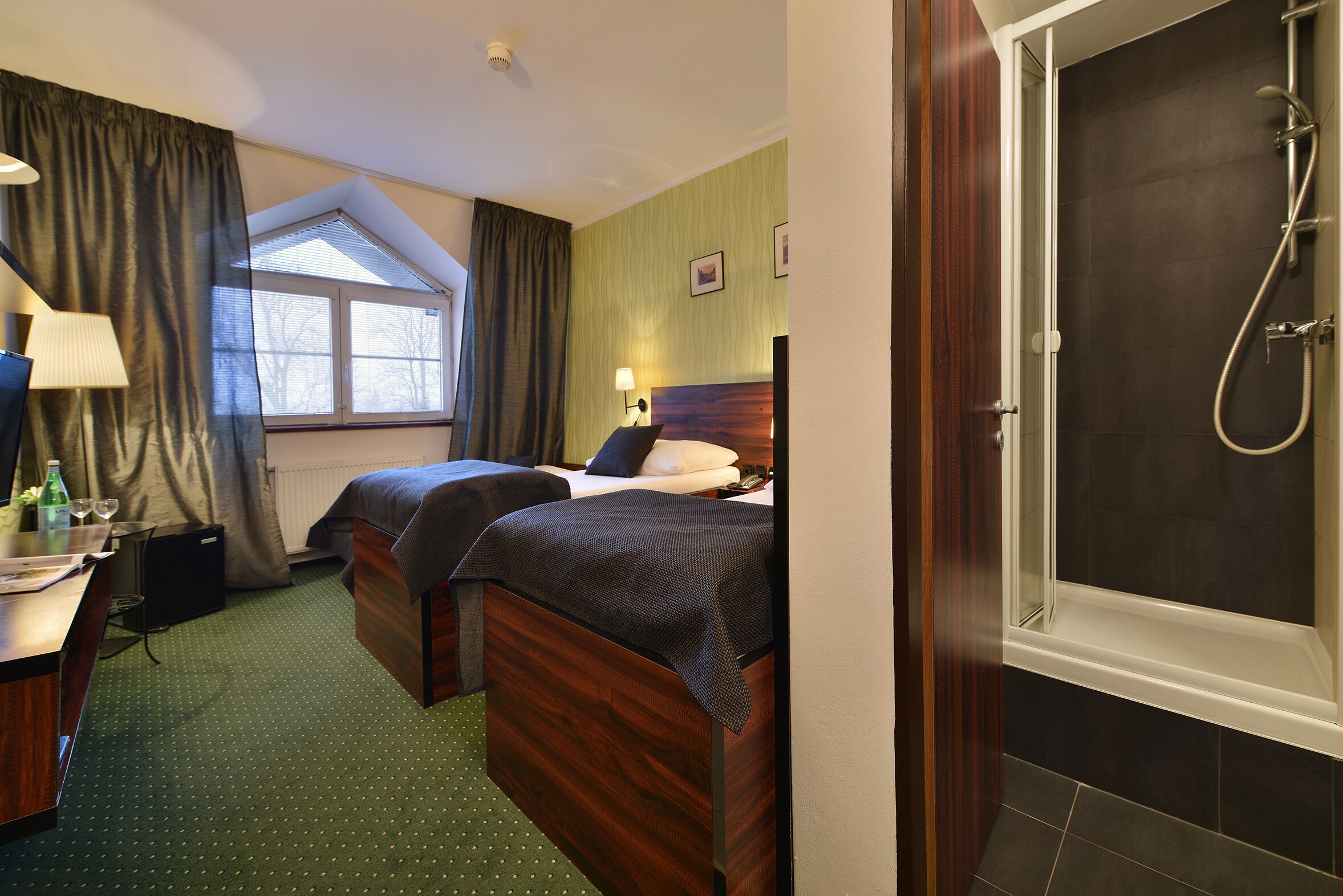 A hotel room with two beds, a desk with a lamp, a window, and a shower area.