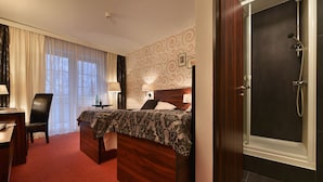 A hotel room with a wooden bed, a desk with a chair, a nightstand, a bathroom with a shower, and a window with curtains. - Parkhotel Morris Nový Bor (Novy Bor)