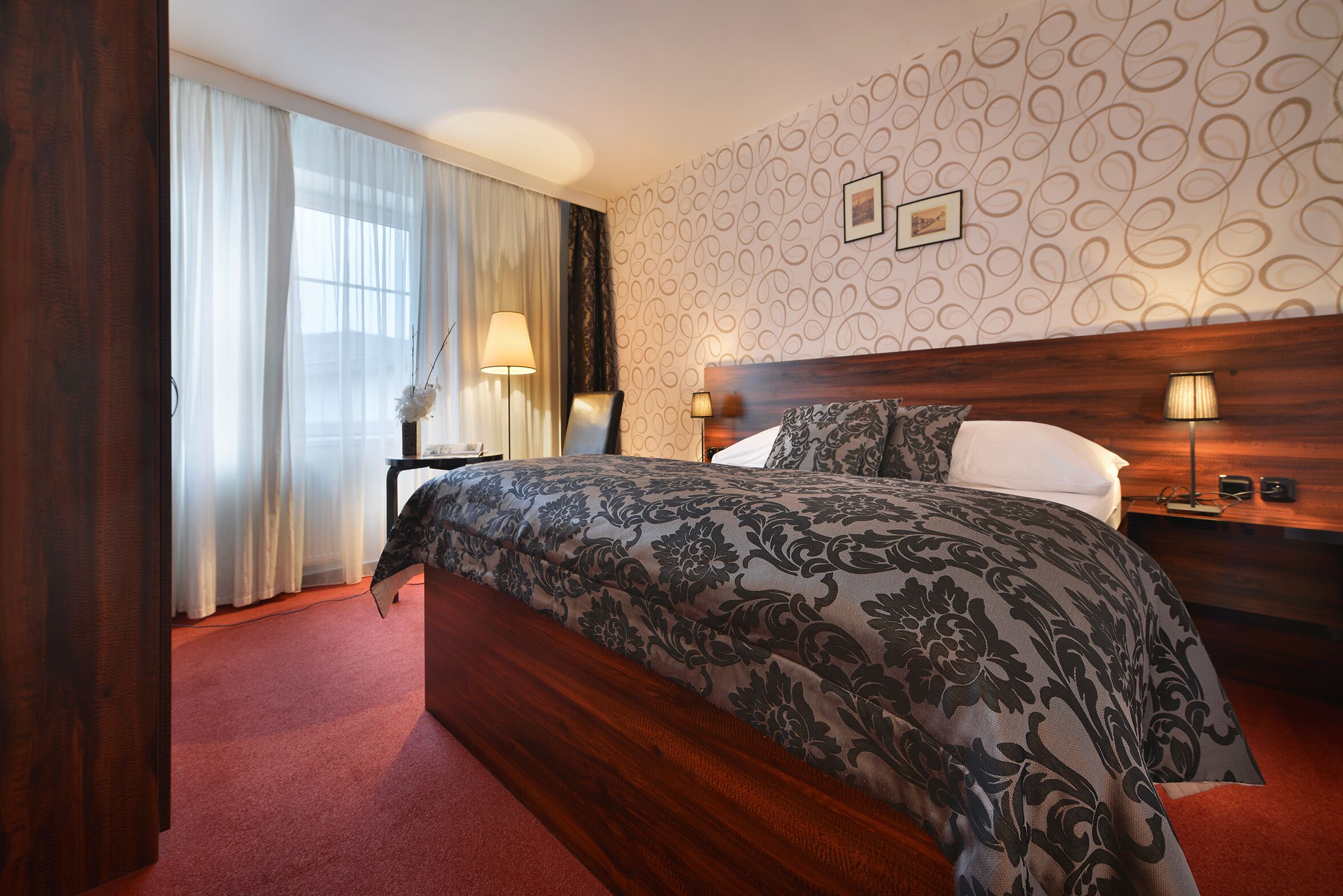 Quarto de hotel com uma cama grande, cabeceira de madeira, parede estampada e janela com cortinas.