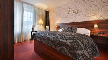 Quarto de hotel com uma cama grande, cabeceira de madeira, parede estampada e janela com cortinas.