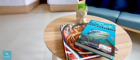 Habitación de hotel con dos camas, un sofá, una mesita con un libro y una planta, y una ventana con cortinas.