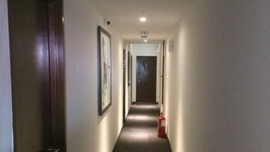 Een smalle, goed verlichte gang met een brandblusser aan de rechterkant, foto’s in de lijstjes aan de muur en een deur aan het einde.