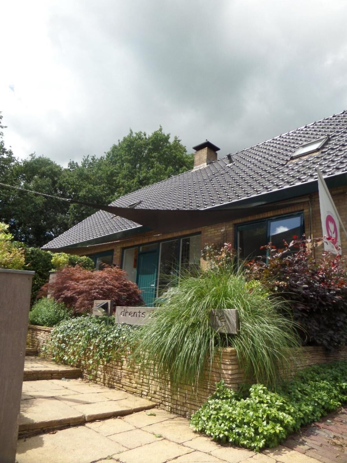A house with a tiled roof, a brick chimney, and a garden with various plants and a sign that reads "drenets".