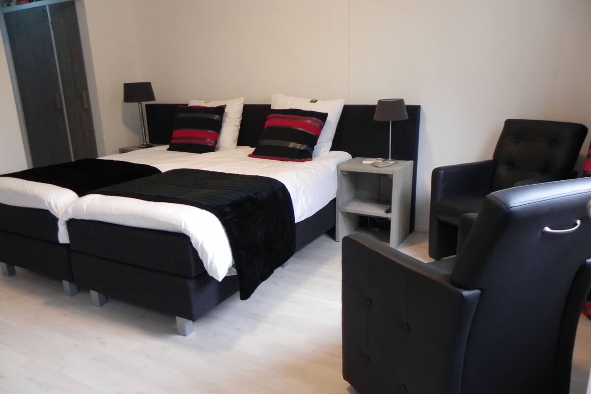 A modern hotel room with a double bed, a side table with a lamp, a black armchair, and a wardrobe.