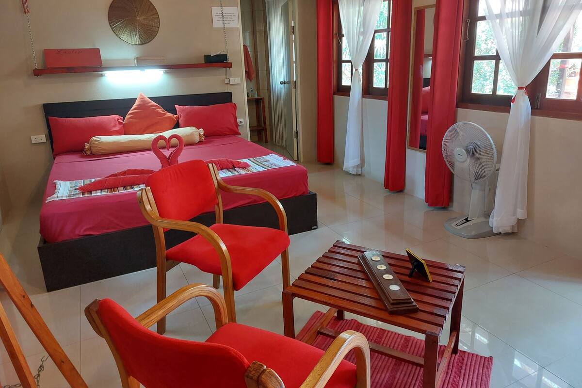 A hotel room with a bed, red chairs, a small table, a fan, and a window with curtains.
